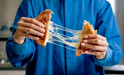 A person in a blue button-down shirt is pulling apart a toasted cheese sandwich, with strings of melted cheese between the two sides.